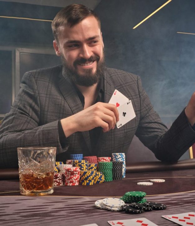 Cheeky brunette man in a classic suit is playing poker sitting at the table at casino in a smoke. He is making bets waiting for a big win. Gambling for money. Games of fortune.