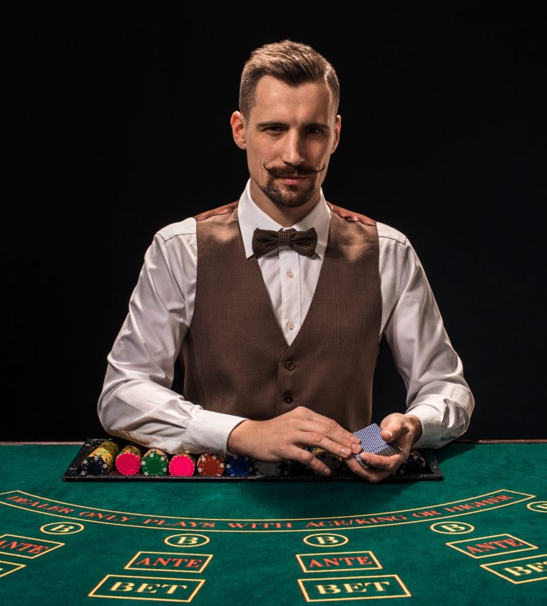 Portrait of a croupier is holding playing cards, gambling chips on table. Black background. A young male croupier in a shirt, waistcoat and bow tie is waiting for you at the blackjack table